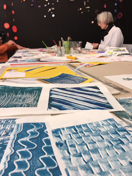 Table de travail avec plusieurs exemples de monotypes sur papier, une personne travaillant en arrière-plan